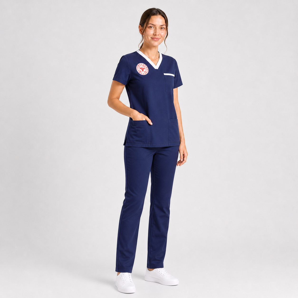 A female healthcare professional standing in a studio setting, wearing a premium navy blue medical scrub set with white v-neck trim details, a red circular chest emblem, and matching straight-leg cargo pants.